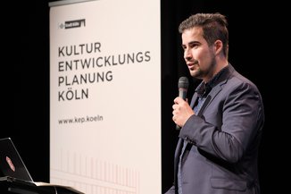 Moderator Sascha Foerster auf der Bühne. Im Hintergrund ein Schriftzug "kulturentwichlungsplanung"